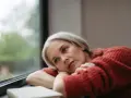 Donna con capelli grigi guarda fuori dalla finestra, pensierosa. Potrebbe cercare rimedi per la depressione estiva.