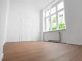Puste, jasne pomieszczenie z białymi ścianami, drzwiami i oknem. Drewniana podłoga i grzejnik sugerują, że to gotowe do zamieszkania mieszkanie socjalne.