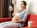 Schwangere Frau mit Halsschmerzen, die sich Sorgen um Scharlach macht. Sie sitzt auf einem roten Sofa.