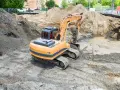 Koparka pracuje na placu budowy, przygotowując teren pod rekultywację. Wokół koparki znajdują się hałdy ziemi i błotniste kałuże.