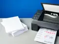 Stos białego papieru obok drukarki, która właśnie drukuje dokument. Różne rodzaje papieru do druku.