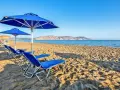 Błękitne leżaki i parasole na piaszczystej plaży Kavros Beach, z widokiem na spokojne morze i górzyste wybrzeże w oddali.