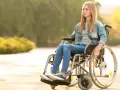 Dziewczyna w dżinsowej kurtce siedzi na wózku inwalidzkim na zewnątrz.