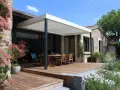 Une belle terrasse tropezienne en bois avec mobilier de jardin sous une pergola moderne. Le soleil brille sur la végétation luxuriante.