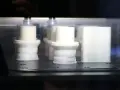 Białe obiekty w drukarce 3D, z widocznym elementem przypominającym 3d print raft, tworzą się warstwa po warstwie.