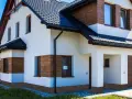 Nowoczesny dom z białym tynkiem i drewnianymi akcentami. Widoczny ciemny dach, okna i drzwi. Dookoła domu biegnie ozdobny cokół.