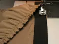 Machine à coudre en train de coudre un élastique sur un tissu beige froncé. Le pied presseur est visible, ainsi que l'aiguille.