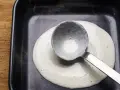 Eine Schöpfkelle verteilt cremige Bechamel-Sauce in einer schwarzen Auflaufform.
