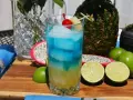 Koktajl z blue curacao alkohol, lodem, wiśnią i ananasem, otoczony owocami: smoczym owocem, limonkami i ananasem.