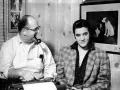 Elvis Presley und Colonel Parker, sein Manager, sitzen nebeneinander. Parker raucht Pfeife, Elvis blickt in die Kamera.