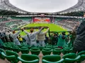 Gwardia Opole kibicuje na stadionie, trzymając szaliki. Na boisku trwa mecz, a trybuny wypełniają fani.