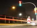 Nachts an einer Kreuzung mit roten Lichtspuren von Autos. Ampeln zeigen Grün und Rot. Ein Vorfahrtsschild und ein Richtungspfeil sind zu sehen. 132 bkat.