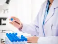 Laborant w białym fartuchu trzyma probówkę z krwią, w tle mikroskop i rząd probówek.
