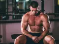 Un homme musclé, concentré, s'entraîne pour prendre du muscle rapidement. Il est assis dans une salle de sport, le regard baissé.