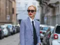 Estilo tomboy que es: mujer con traje a cuadros, camisa blanca y corbata negra, gafas de sol y labios rojos, posando en la calle.