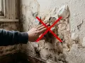 Mano tocando una pared bufada con moho y pintura descascarada. Una cruz roja indica que no se debe tocar.