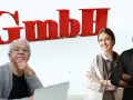 Drei Personen vor rotem "GmbH"-Schriftzug. Ein älterer Mann sitzt am Laptop, eine junge Frau und ein Mann stehen lächelnd daneben. Sie repräsentieren die GmbH-Konzession.