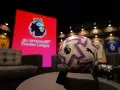 Piłka do gry w Premier League na tle ekranu z logo ligi. W tle widoczne koszulki piłkarskie.