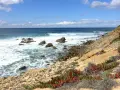 Fischerweg Portugal: Felsenküste mit Wellen, blauem Himmel und Wolken. Sukkulenten wachsen am Hang.