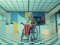 Zofia Komasa w wózku inwalidzkim, otoczona nowoczesnymi ekspozycjami, z kolorowym tłem i charakterystyczną podłogą w szachownicę.