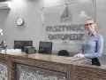 Uśmiechnięta recepcjonistka w okularach za ladą w gabinecie ortopedycznym.