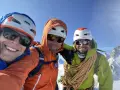 Trois alpinistes souriants au sommet, prêts à devenir guide de haute montagne à 40 ans. Corde et équipement de sécurité visibles.