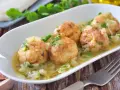 Cuatro albóndigas de merluza doradas en una salsa cremosa con cebolla y perejil, servidas en un plato blanco.