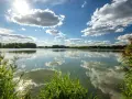 Sonnenschein über dem celle see, mit Wolken, die sich im ruhigen Wasser spiegeln. Schilf säumt das Ufer.