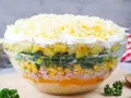Schichtsalat-Rezept in einer Glasschüssel: Schichten aus Ananas, Lauch, Mais, Schinken, Orange, Ei und Käse.