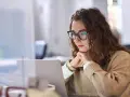Donna con occhiali lavora al laptop, pensierosa. Potrebbe essere una dei nomadi digitali chi sono, che lavora da remoto.