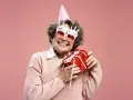 Uśmiechnięta starsza pani w okularach z napisem "Happy Birthday" i w czapce urodzinowej trzyma prezent. To dowcipne podziękowania za życzenia!