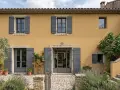 Façade d'une maison provençale à l'enduit à la chaux ocre, avec volets gris et végétation luxuriante.