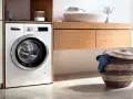 Lavadora blanca moderna lista para instalar en un baño con cesta de ropa sucia.