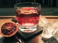 Ein Glas mit einem roten Drink, garniert mit Orangenschale, neben einer Blutorange und Eiswürfeln. Das ist Campari, ein italienischer Aperitif.
