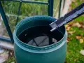 Zielona beczka zbiera deszczówkę z rynny. W tle szklarnia i jesienne liście.