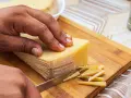 Mano cortando un bloque de queso amarillo en finas láminas. La corteza del queso se ve firme, indicando que se puede comer.