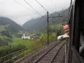 A smiling man leans out the window of the Gotthard Panorama Express, enjoying the stunning Swiss Alps scenery with a church nestled in the valley below.