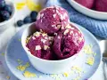 Drei Kugeln Blaubeer Eis, garniert mit weißen Schokostückchen, in einer weißen Schale auf einem blauen Teller.