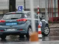 Samochód egzaminacyjny z literą "L" na dachu i napisem "EGZAMIN" na tylnej klapie, otoczony pachołkami na mokrym placu.