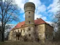 Zniszczony pałac w Osetnie z charakterystyczną wieżą i czerwoną dachówką, otoczony drzewami.
