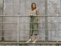 Kobieta w zielonym kombinezonie i białych szpilkach stoi przy metalowej balustradzie na tle betonowej ściany.