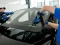 Dwóch mechaników wymienia przednią **szybę** w czarnym samochodzie, używając przyssawek.