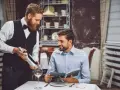 Camarero atento presenta una botella de vino a un cliente que revisa el menú. Demuestra las cualidades de un camarero: servicio, amabilidad y conocimiento.