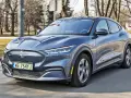 Niebieski Ford Mustang Mach-E jedzie po drodze, z widocznym logo konia na grillu.