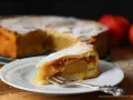 Ein Stück von einem köstlichen, mit Puderzucker bestäubten gedeckten Apfelkuchen liegt auf einem Teller. Daneben ein ganzer Kuchen und Äpfel.
