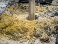 Wiertnica zagłębia się w ziemię, tworząc otwór pod fundamenty palowe. Wokół leżą hałdy ziemi i kamieni.