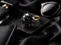 Elegancki pierścionek z czarnym kamieniem szlachetnym, obok flakonika perfum i okularów. Czarne kamienie szlachetne nazwy: diament, spinel, turmalin.