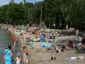 Tłoczna plaża nad jeziorem na Pomorzu. Ludzie opalają się, kąpią i odpoczywają w cieniu drzew.