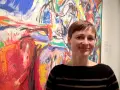 Kvinna ler framför en färgstark, abstrakt målning i Asger Jorns anda.