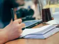 Mano escribiendo en un cuaderno abierto, inspirada para escribir cuentos. En la mesa hay una tableta, una taza y una jarra.